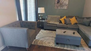 Ein Wohnzimmer mit grauem Sessel, einer gelb gekissenen Couch, einem gemusterten Teppich und einem Bild an der Wand.
