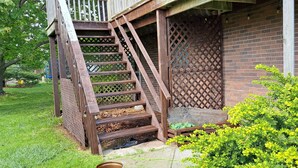 Un escalier extérieur en bois menant à une véranda agrémentée d’un treillis décoratif, le tout entouré de verdure et d’un mur de briques.