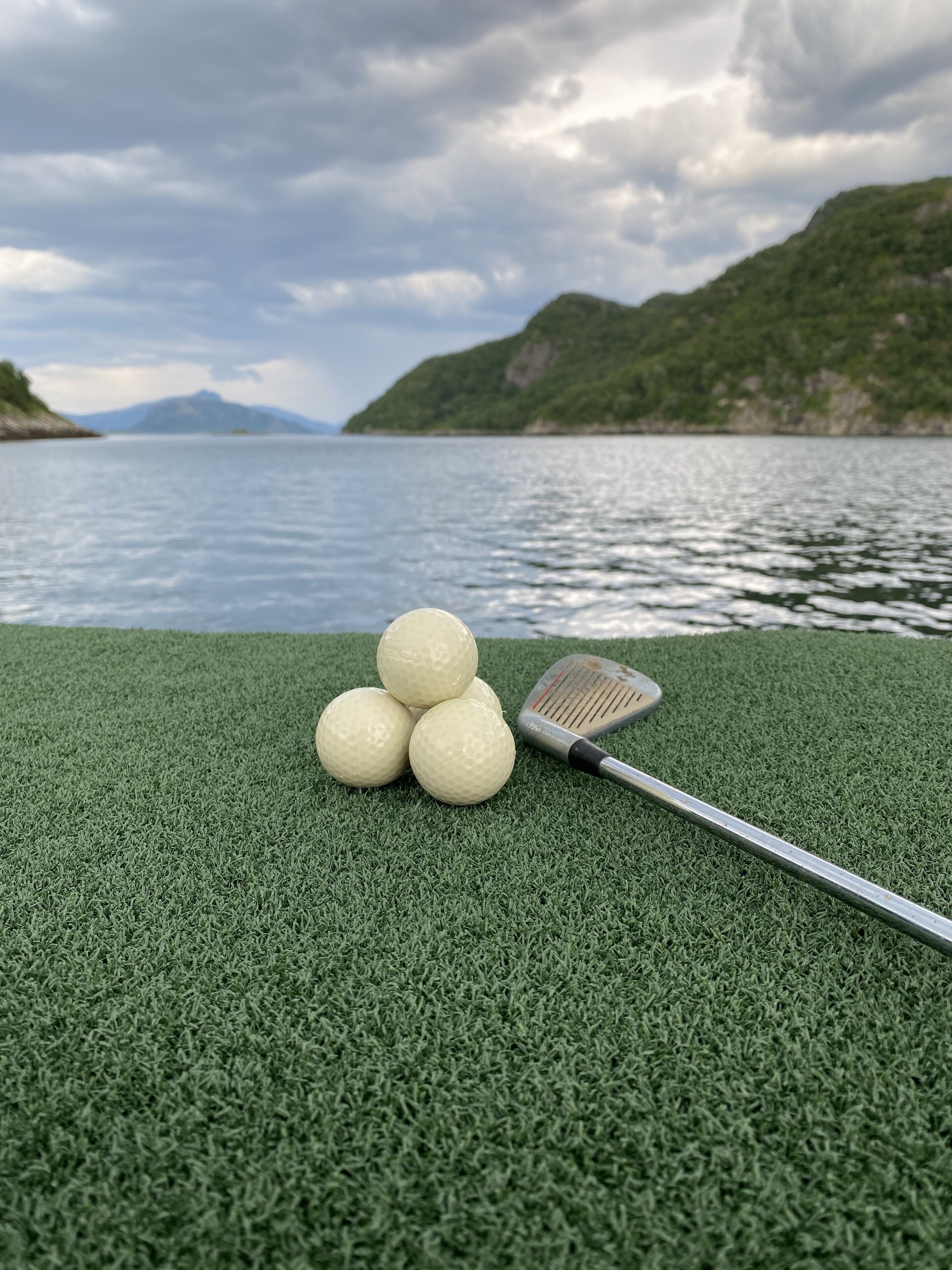 Bolas de golfe e taco sobre um campo verde, perto de um corpo de água, com montanhas ao fundo.