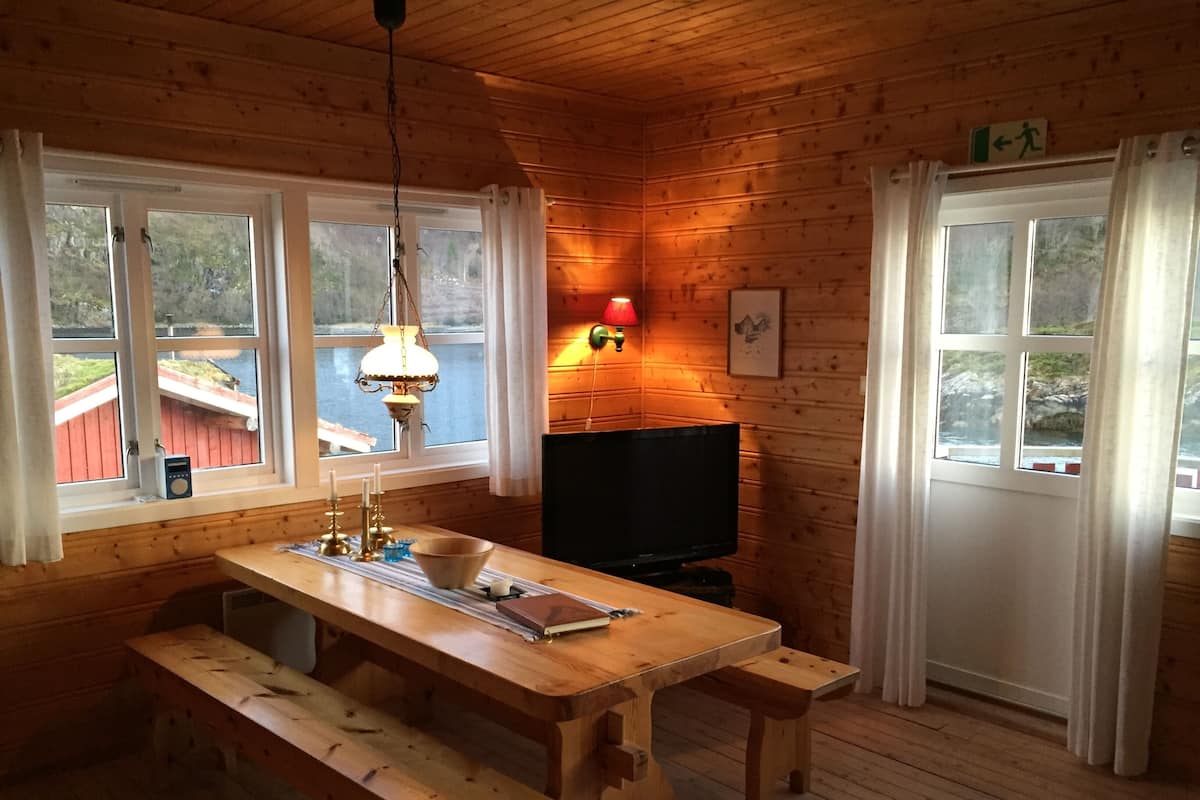 A cozy cabin interior with a wooden dining table, benches, a television, and large windows offering a view of a dock and water.