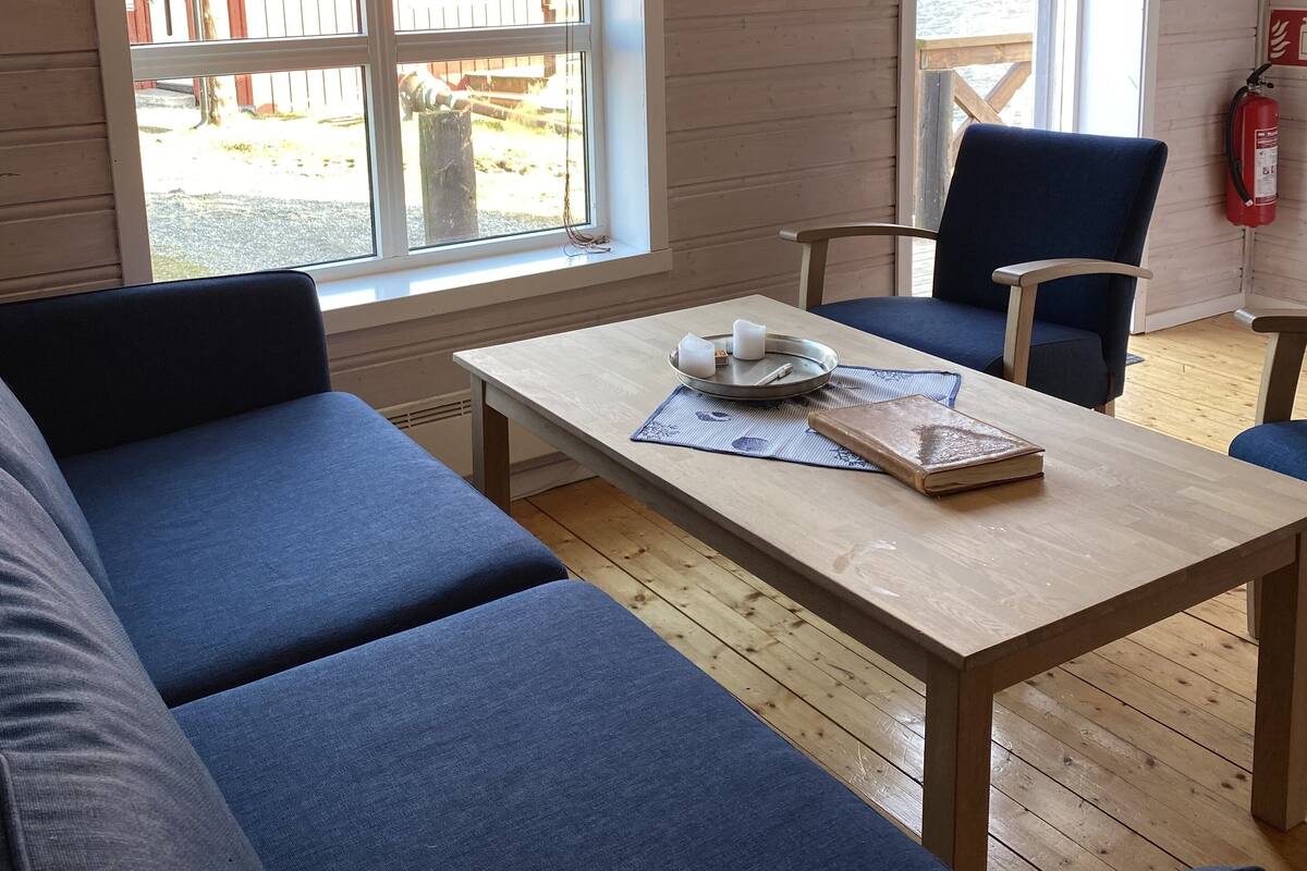 A room with a wooden floor, a sofa, a table with a book and candles, and a window with a view of a wooden structure outside.