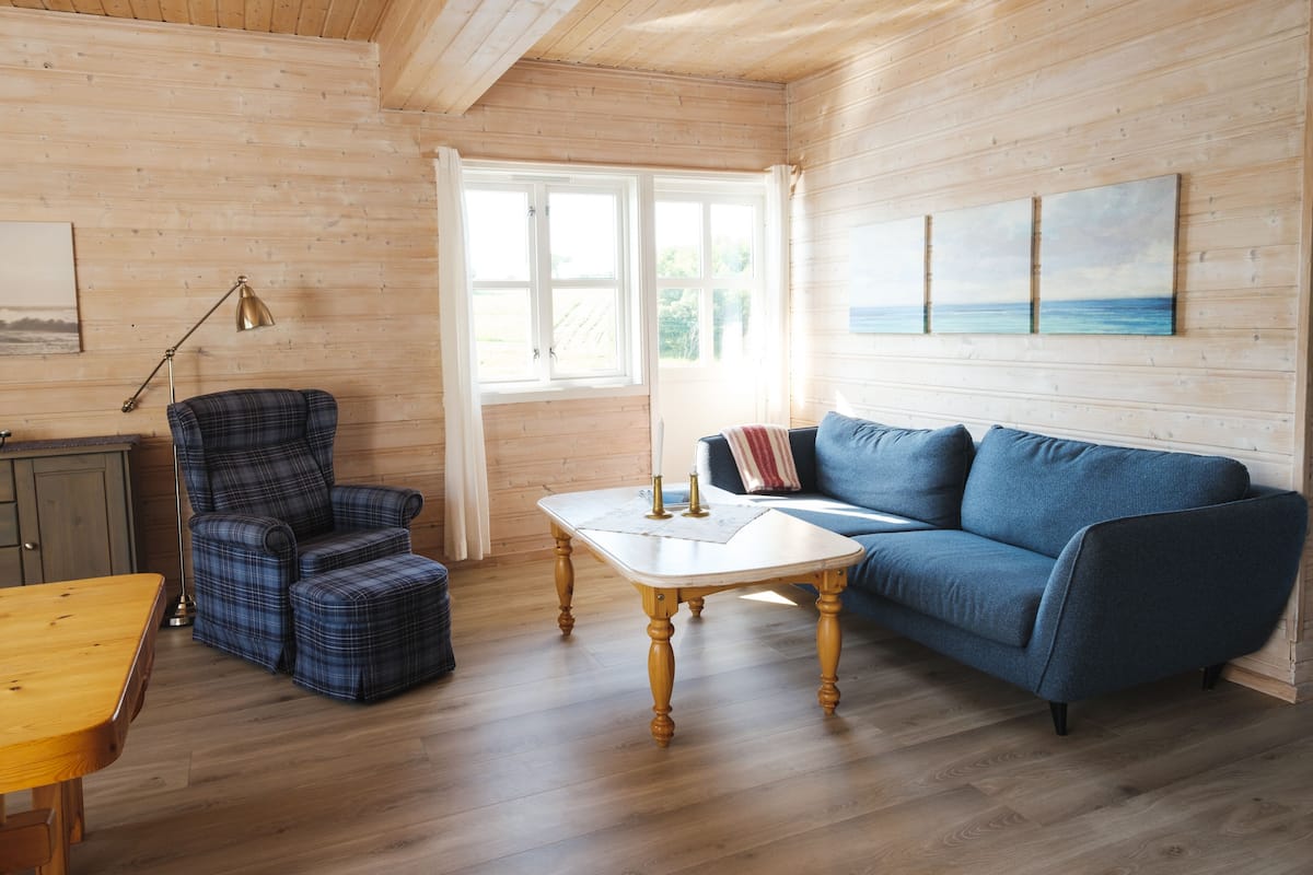 A cozy room with a blue sofa, plaid armchair, wooden coffee table, and a view of the outdoors through a large window.