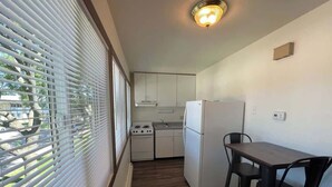 A compact kitchen with white appliances, a dining table with chairs, and a window with blinds. - Liberty Valley Studios in Madison, Wi! (Madison)