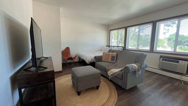 A modern living room with a flat-screen TV, a grey sofa, a small ottoman, and a wicker rug. - Liberty Valley Studios in Madison, Wi! (Madison)