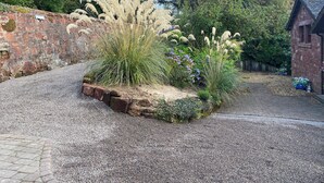 Een grindpad dat leidt naar een rustiek stenen gebouw met een bakstenen muur en een tuin met hoge grassen en bloeiende planten.