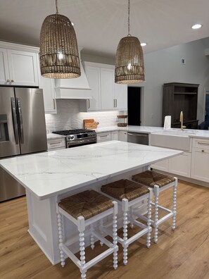 A modern kitchen with white cabinetry, a large island with stools, stainless steel appliances, and wicker pendant lighting. - FRIPP ISLAND/Hot tub overlooking lake/ Community Pool/6 Seater Golf Cart/Cards available-275 Deer Lake (Fripp Island)