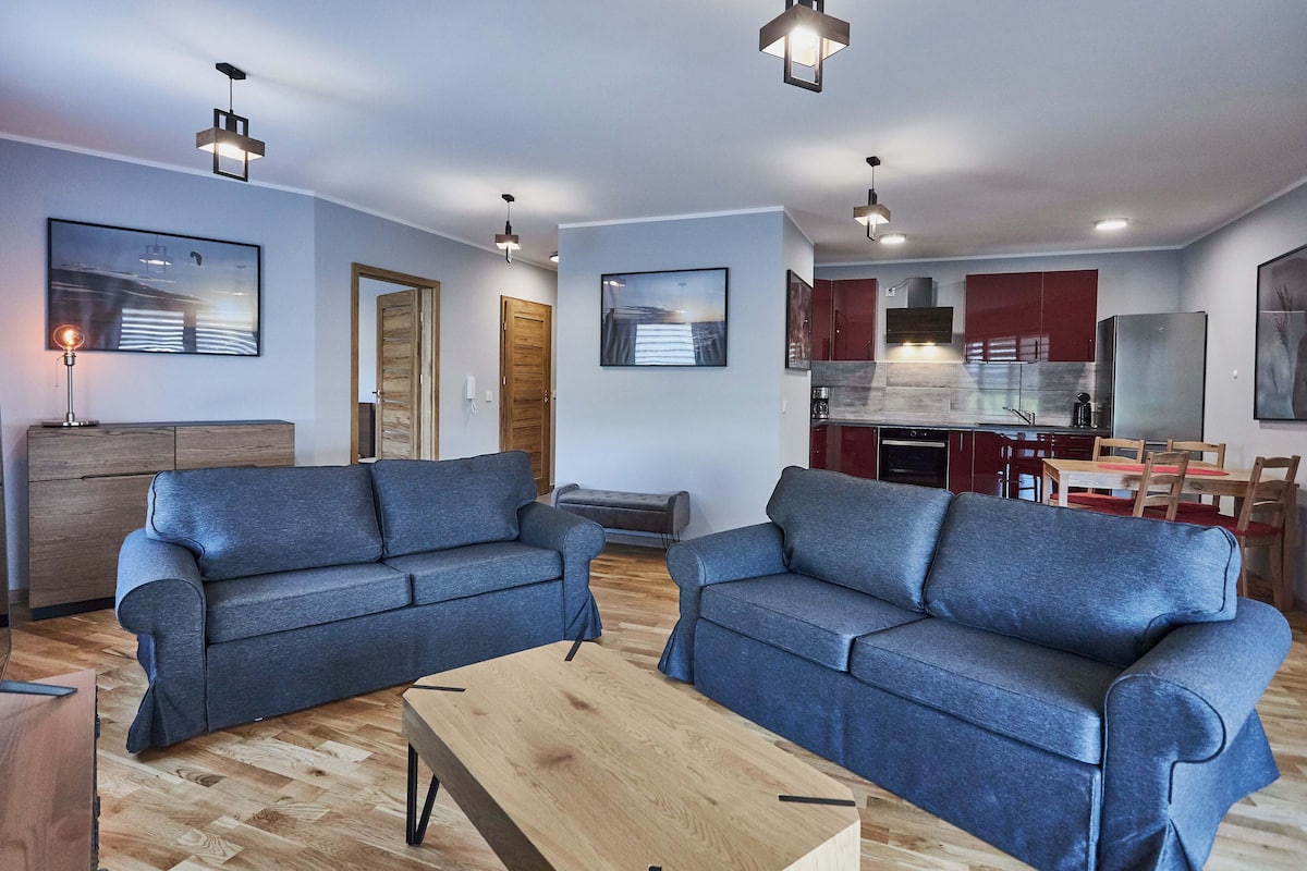 A modern living room with two grey sofas, a wooden coffee table, and a kitchen area with red cabinets and stainless steel appliances.