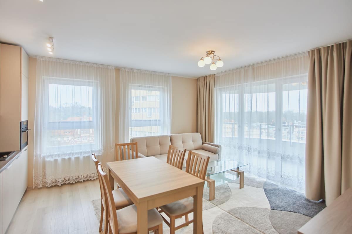 A modern living room with a sofa, dining table, and chairs. There are large windows with sheer curtains and a view of buildings outside.