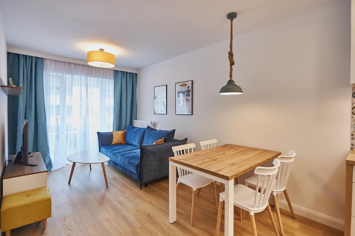 A modern living room with a blue sofa, a wooden dining table with chairs, a standing lamp, and a large window with curtains.