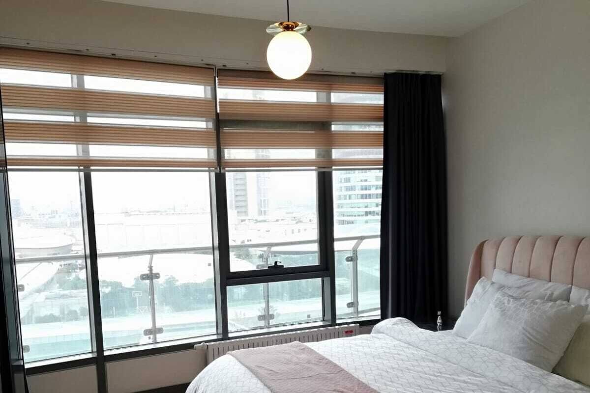 Ein modernes Schlafzimmer mit einem großen Bett, Blick auf die Stadt durch raumhohe Fenster und einer Pendelleuchte.
