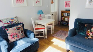 A living room with a blue armchair, a white table, and a blue sofa. There are framed pictures on the wall, a lamp on the table, and a bookshelf in the background. - Holmlea Cottage, a sweet and charming getaway in a picturesque Fife village. (Freuchie, Fife)