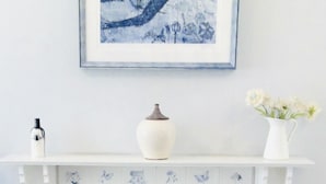 A white fireplace mantel with a framed blue artwork, a white vase with flowers, and a white vase with a brown lid. - Holmlea Cottage, a sweet and charming getaway in a picturesque Fife village. (Freuchie, Fife)