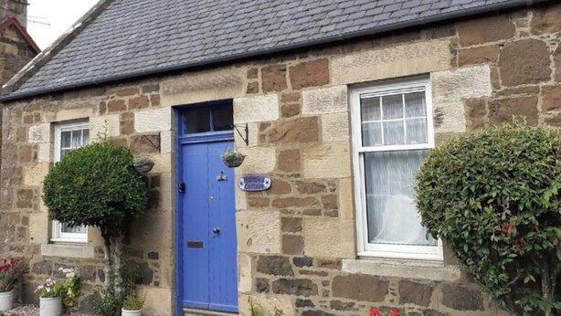 A stone cottage with a blue door, a small garden, and a sign indicating it is a holiday cottage. - Holmlea Cottage, a sweet and charming getaway in a picturesque Fife village. (Freuchie, Fife)