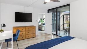 A modern bedroom with a ceiling fan, a TV, a wooden dresser, a desk, and a blue bedspread. - Loggerhead 351 | Sunny Sanibel | Gorgeous Walkout Unit | Gulf Views (Sanibel)
