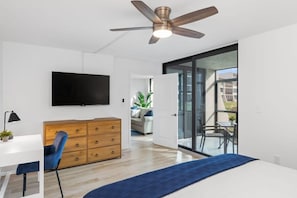 A modern bedroom with a ceiling fan, a TV, a wooden dresser, a desk, and a blue bedspread. - Sunny Sanibel - Gorgeous Walkout Unit at Loggerhead Cay Gulf Views (Sanibel)
