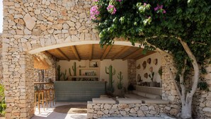 Un espacio al aire libre con paredes de piedra, un banco de madera, un jardín de cactus y una zona de estar con un banco de piedra y cojines.