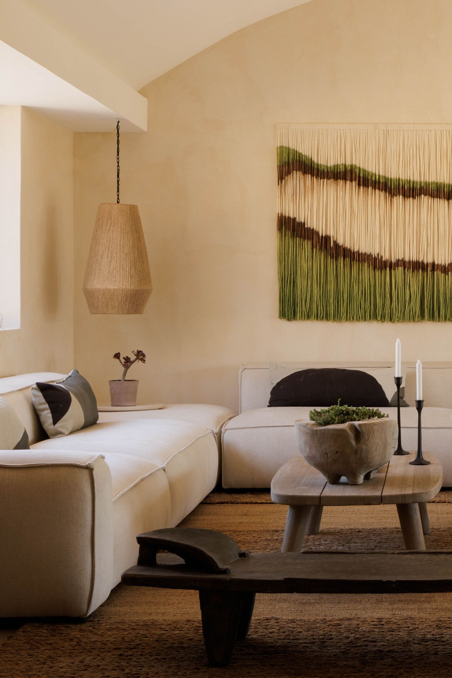Un salon moderne avec un canapé d’angle, un luminaire suspendu, une tapisserie murale et une petite table avec des bougies.