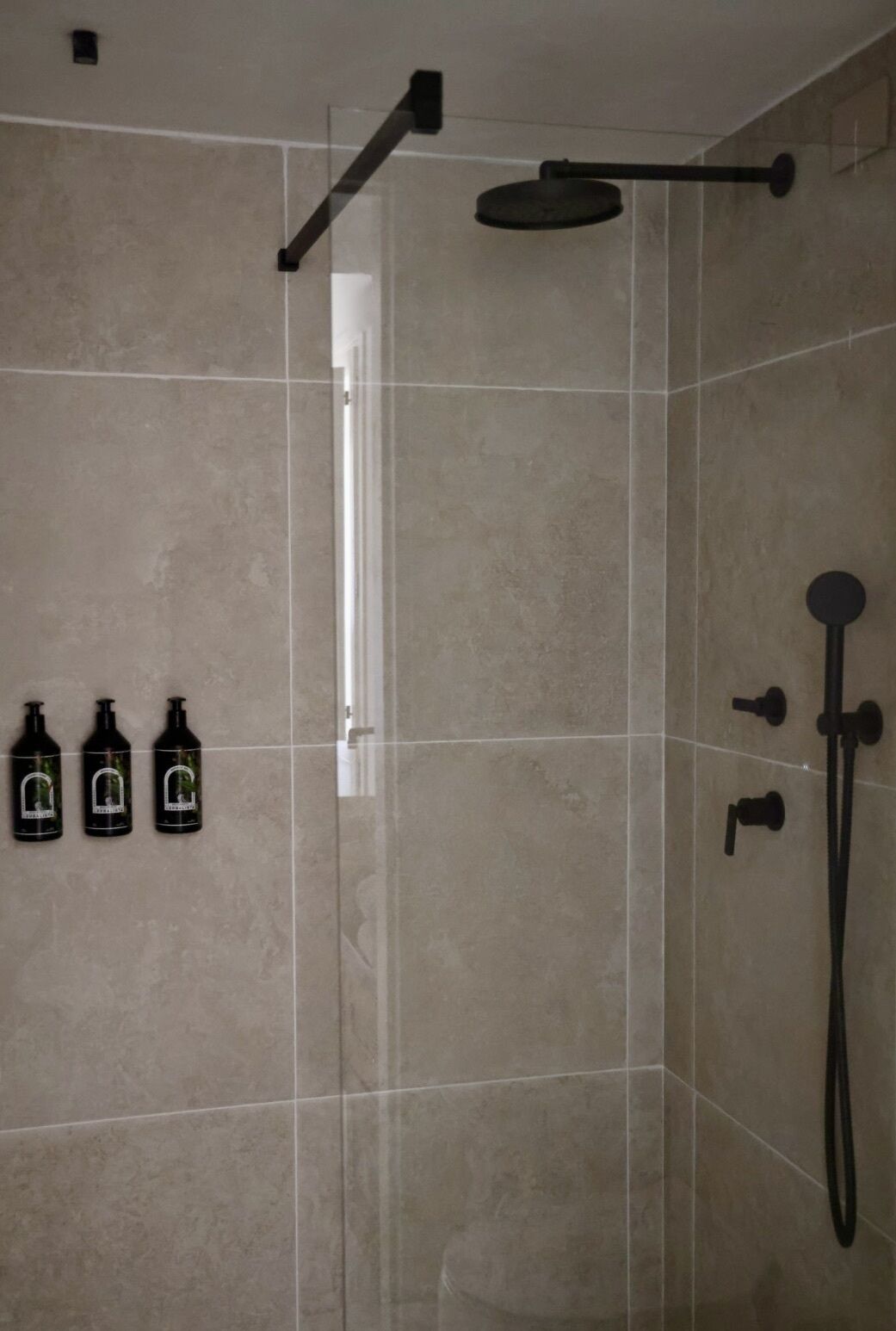 A shower area with a glass door, a handheld showerhead, and three bottles of shampoo on the wall.