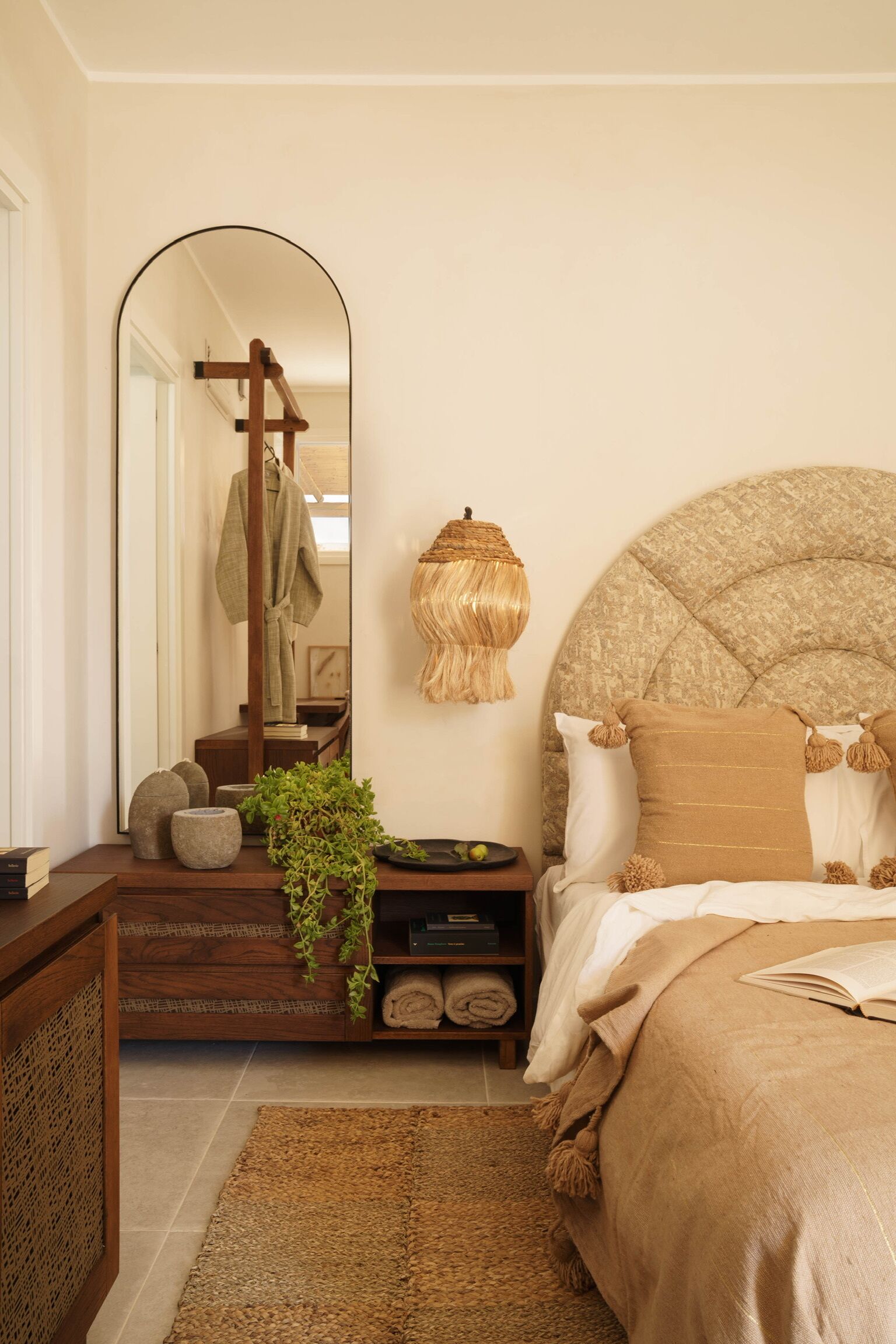 A bedroom with a large mirror, a round headboard, a bedside table with a plant, and a bed with a brown blanket.