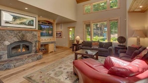 A spacious living room with a stone fireplace, leather sofas, and large windows offering a view of the outdoors. - Lazy Bear Lodge in Homewood - hot tub, close to lake, large home (Homewood)