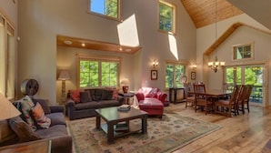A spacious living room with a high ceiling, large windows, and a dining area with wooden furniture. - Lazy Bear Lodge in Homewood - hot tub, close to lake, large home (Homewood)