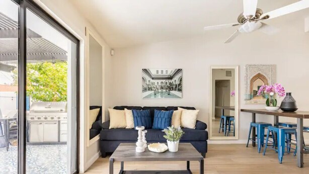 A modern living room with a black sofa, a coffee table, and a dining area with blue stools. -  Desert Escape w/Pool/Jacuzzi (Palm Desert)