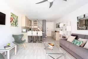 Ein modernes Wohnzimmer mit einem grauen Sofa, einem Glastisch und einem grünen Sessel. Es gibt eine Küchenzeile mit Esstisch und eine Treppe, die zu einer oberen Ebene führt.