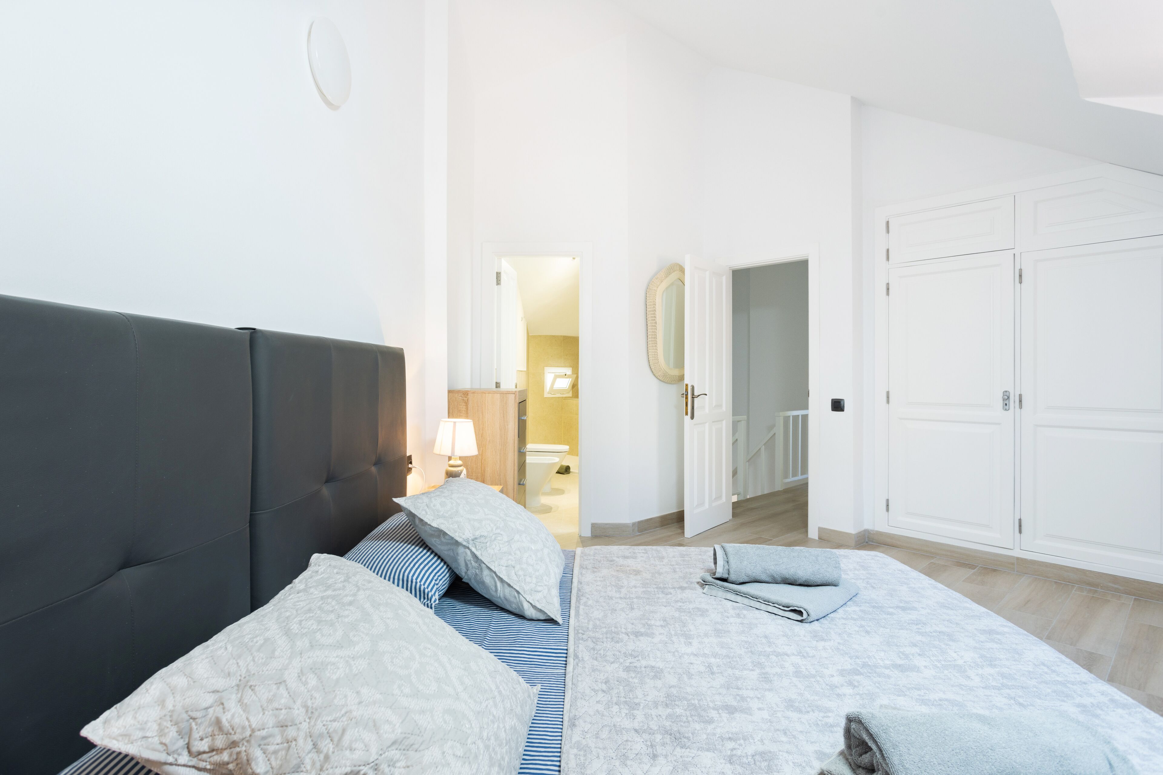 Ein Schlafzimmer mit dunklem Kopfteil, einem Bett mit gemusterten Betttextilien, einer Nachttischlampe sowie Blickrichtung auf ein Badezimmer und einen Flur.