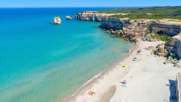 Uma praia costeira com águas turquesa cristalinas, falésias rochosas e uma faixa de areia com guarda-sóis e cadeiras de praia.