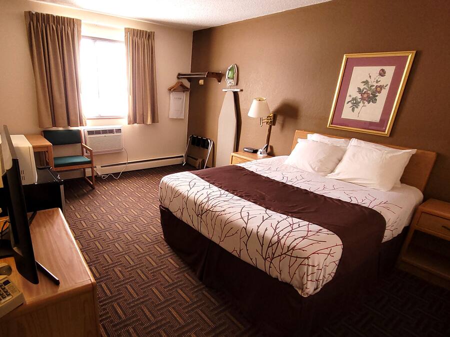 A hotel room with a large bed, a desk with a computer, a chair, an air conditioning unit, a lamp, and a framed picture on the wall.