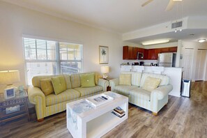 A living room with a striped sofa, a coffee table, and a view of the kitchen. - Private Waterfront Balcony! Steps From the Beach, Heated Pool, with Nearby Grocery & Restaurants. (Treasure Island)