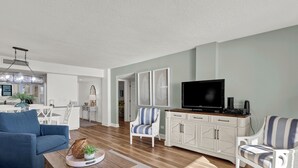 A modern living room with a TV, white furniture, and a wooden floor. - Station One 6F - a beautifully updated 2 bedroom condo (Wrightsville Beach)