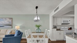 A modern living room with a blue armchair, a dining table with white chairs, a painting on the wall, and a kitchen area with white cabinets and appliances. - Station One 6F - a beautifully updated 2 bedroom condo (Wrightsville Beach)