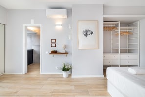 A modern bedroom with a wooden floor, a white bed, a walk-in closet with hanging space, and a small plant on the wall.