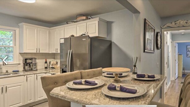 Una cocina moderna con armarios blancos, un refrigerador de acero inoxidable y una zona de comedor con encimera de mármol y una mesa redonda.