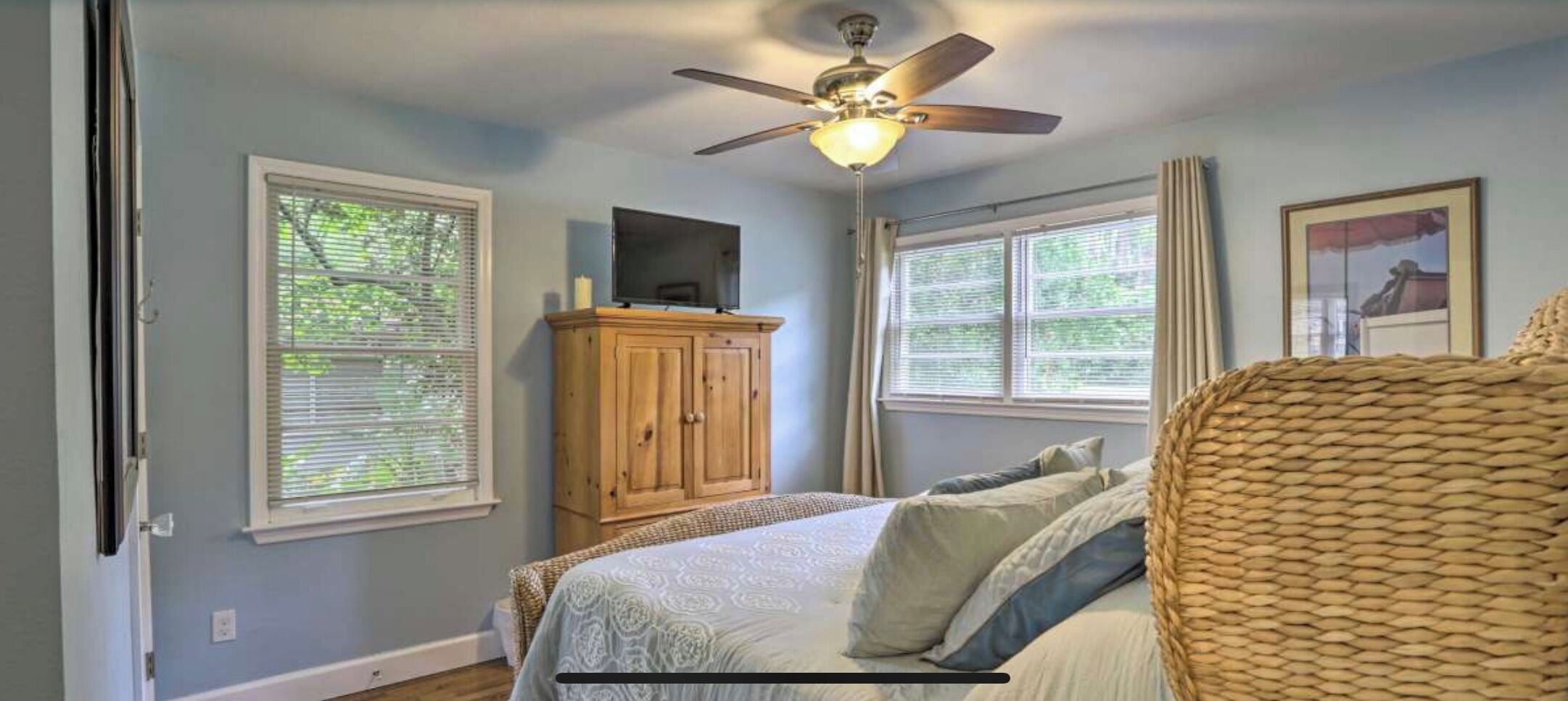 Une chambre à coucher comprenant un lit, un ventilateur de plafond, une armoire en bois et deux fenêtres équipées de stores.