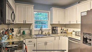 Una cocina con armarios blancos, un refrigerador de acero inoxidable, una ventana con vista a árboles y diversos electrodomésticos.