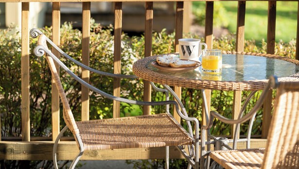 Een rieten tafel met een glas sinaasappelsap en een mok, op een houten terras met stoelen.