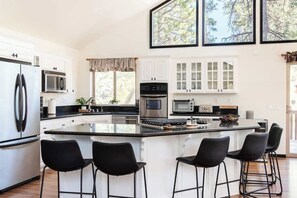 Une cuisine moderne avec des meubles blancs, un îlot central doté de tabourets et de grandes fenêtres qui laissent entrer la lumière naturelle.