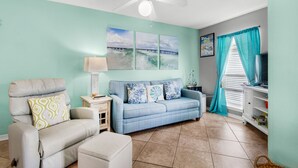 A living room with a teal wall, a white ceiling fan, a blue sofa, a white armchair, a small table with a lamp, and a TV on a white cabinet. - 🌅Steps2Sound|Pool|Spacious 1BR|Sunset Harbor Villas 313🌅 (Navarre Beach)