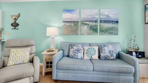 A living room with a teal wall, a gray sofa, and a matching armchair. There are three wall-mounted paintings depicting a bridge and waves. - 🌅Steps2Sound|Pool|Spacious 1BR|Sunset Harbor Villas 313🌅 (Navarre Beach)