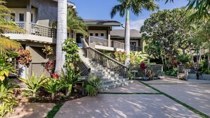 A multi-story building with a staircase leading to a balcony, surrounded by tropical plants and palm trees.