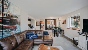 A modern living room with a brown leather sofa, a coffee table, and a painting of a boat on the wall. There is a kitchen in the background with wooden cabinets and a dining area with stools. - Baker View Lodging with hot tub, sound, mountain, and territorial views. (Camano)