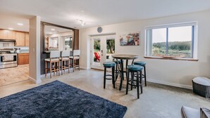 A modern kitchen with wooden cabinets, a dining area with a round table and bar stools, and a large window with a view of greenery. - Baker View Lodging with hot tub, sound, mountain, and territorial views. (Camano)
