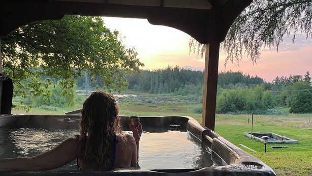 A person in a hot tub with a view of a forest and a sunset. - Baker View Lodging with hot tub, sound, mountain, and territorial views. (Camano)