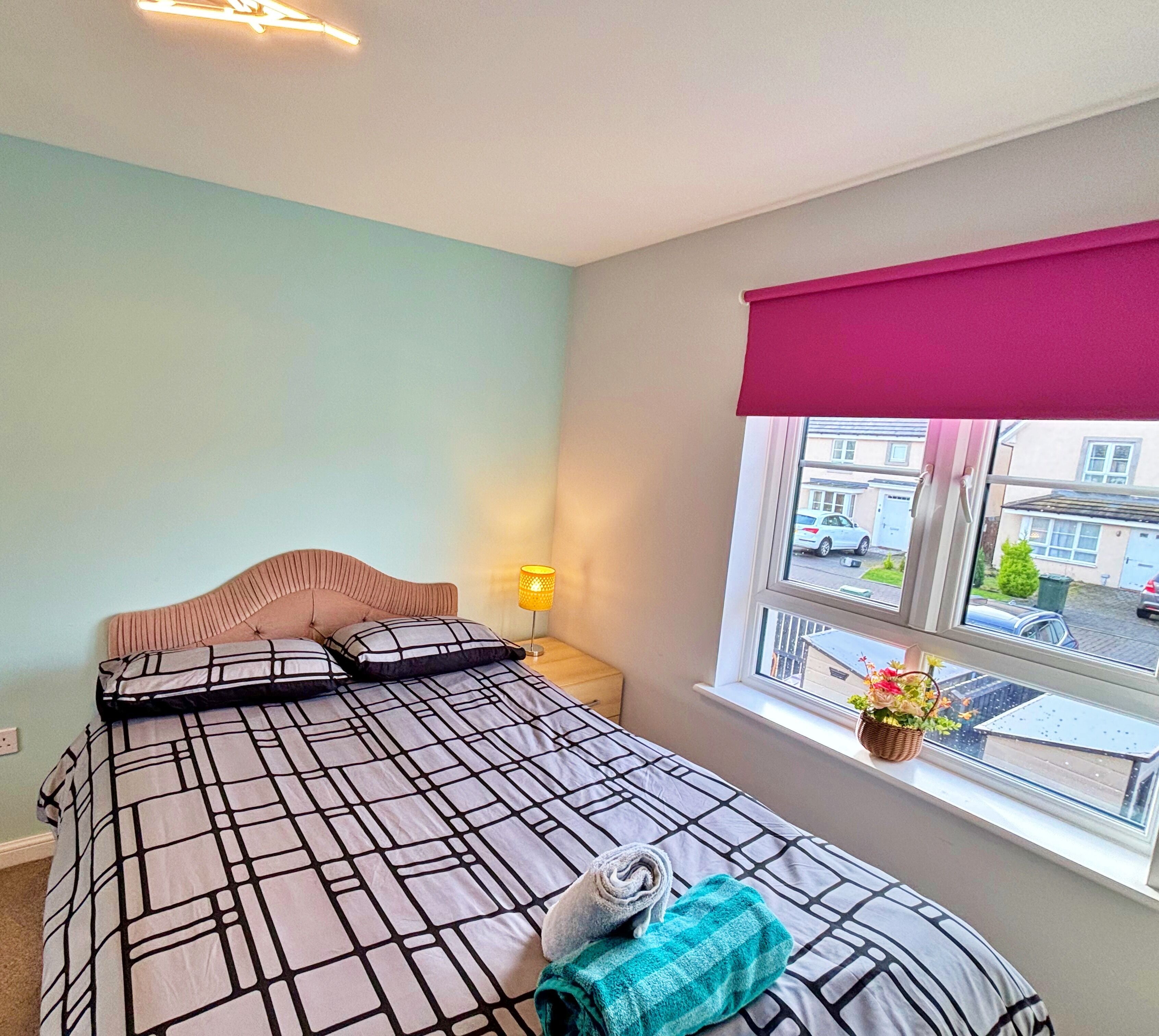 A bedroom with a bed, a window with a view of the street, and a nightstand with a lamp.