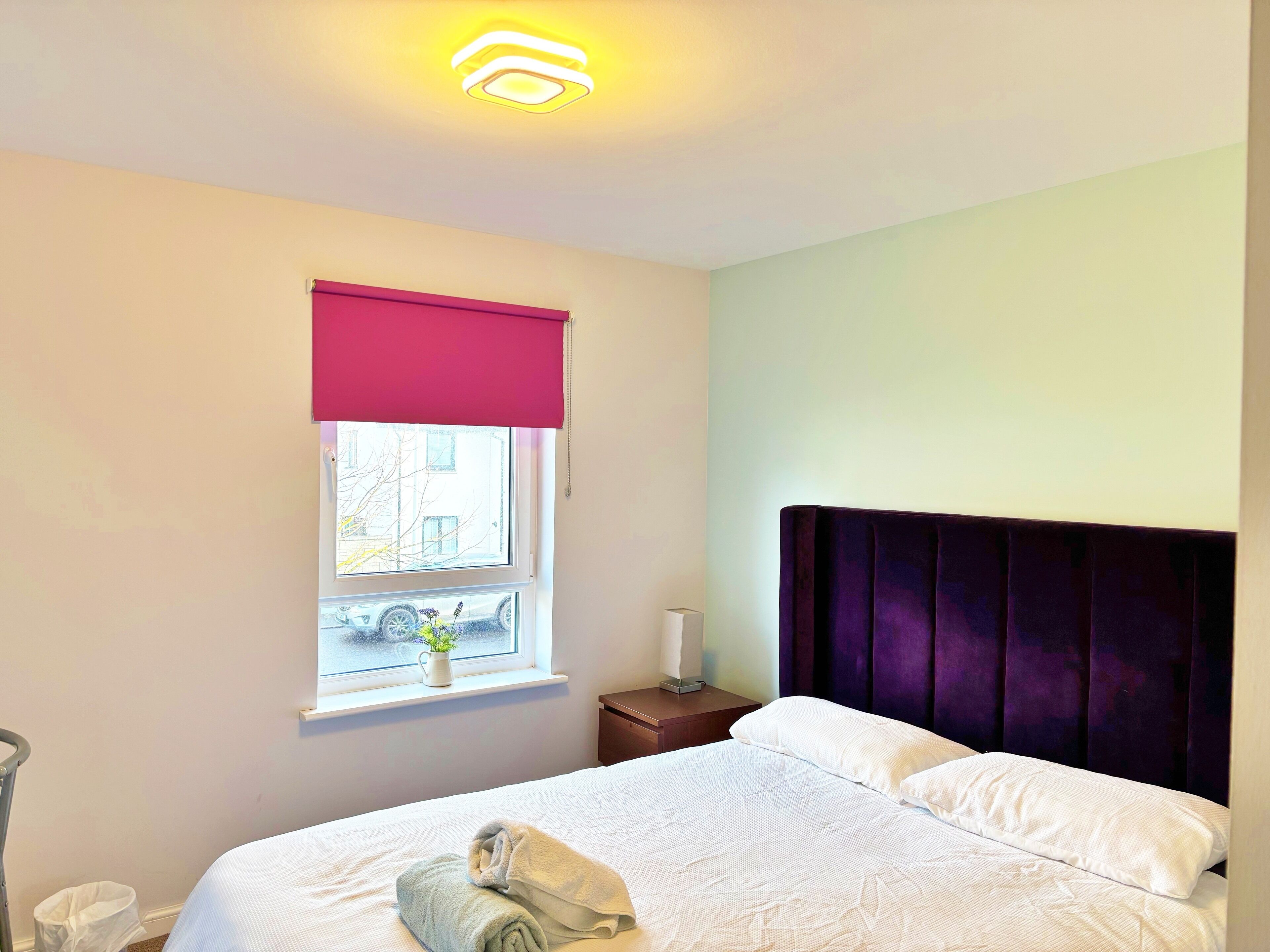 Un lit bien fait, avec du linge de lit blanc, une fenêtre avec un store rose et une table de chevet avec une lampe.