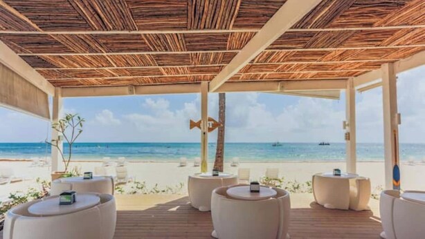 Een strandlounge met een houten plafond, witte, beklede zitplaatsen en uitzicht op zee.
