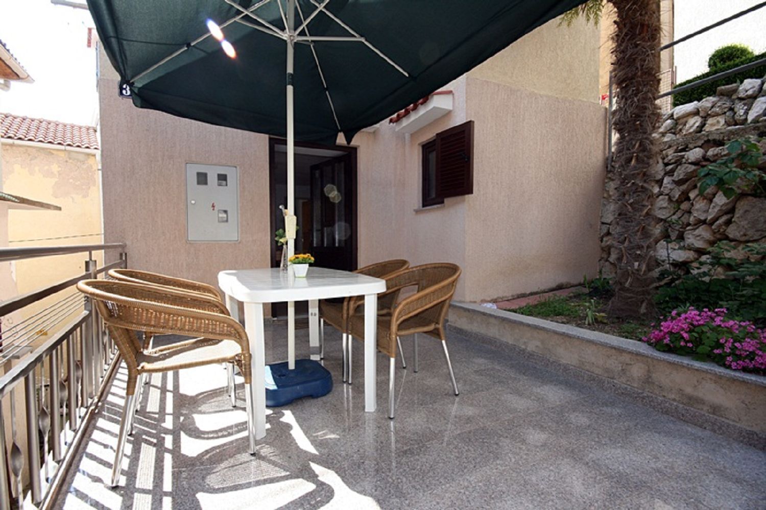 Eine Terrassenanlage im Freien mit Tisch, Stühlen, einem grünen Sonnenschirm und einer Steinmauer mit Pflanzen.