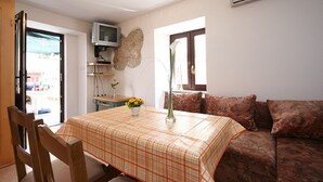 A dining area with a table set for two, a television mounted on the wall, and a sofa with patterned cushions. - 100 metres to the pebble beach (Baska)